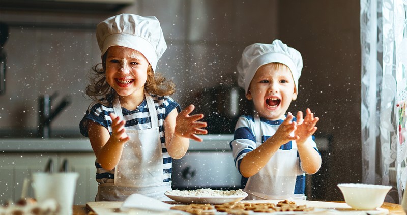A photo of two children in a kitchen, dressed in chefs whites, smiling as they bake. There is flour that can be seen in the air. 