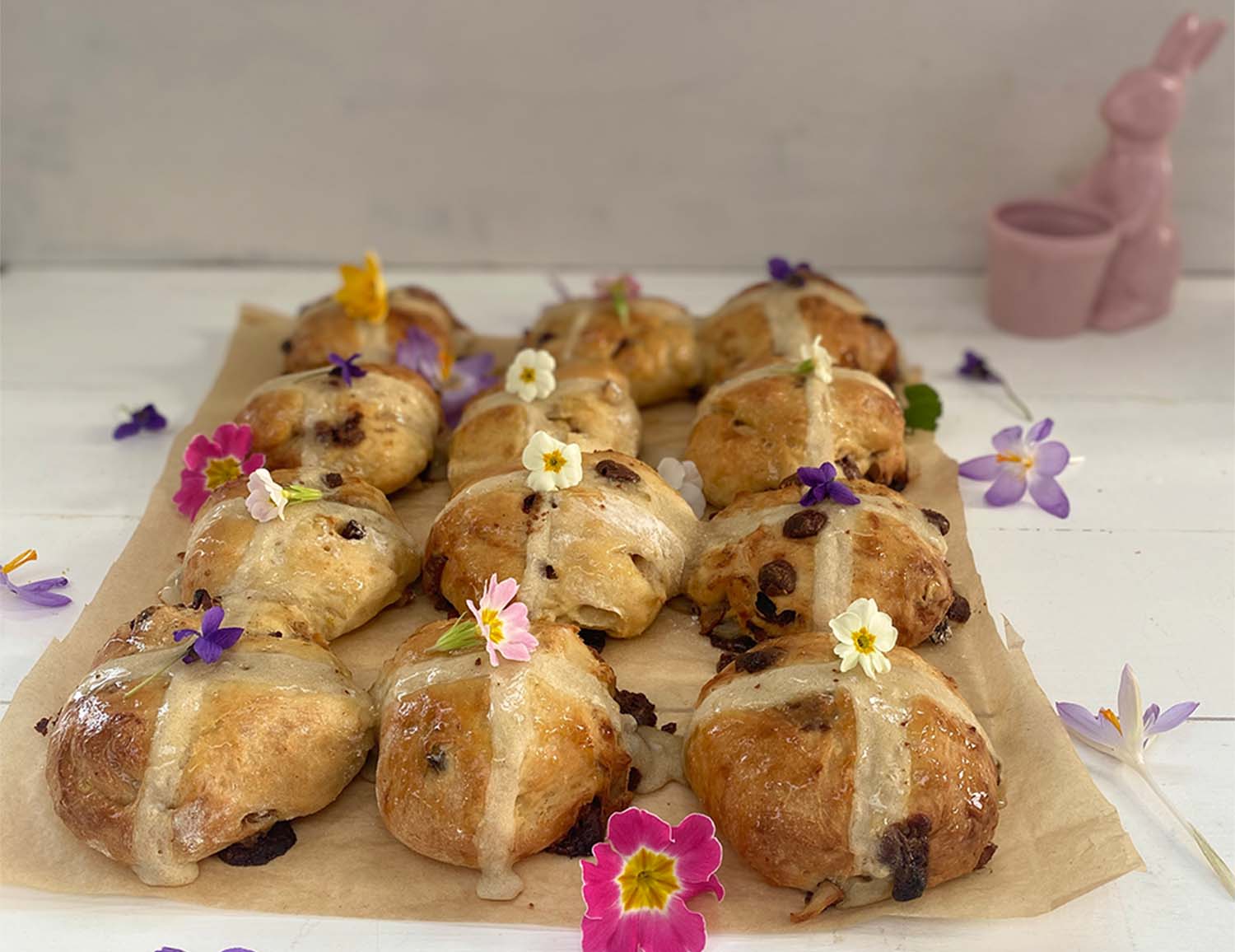 An image of nine milk chocolate, pear and honey hot cross buns, set on baking paper and decorated with flowers. 