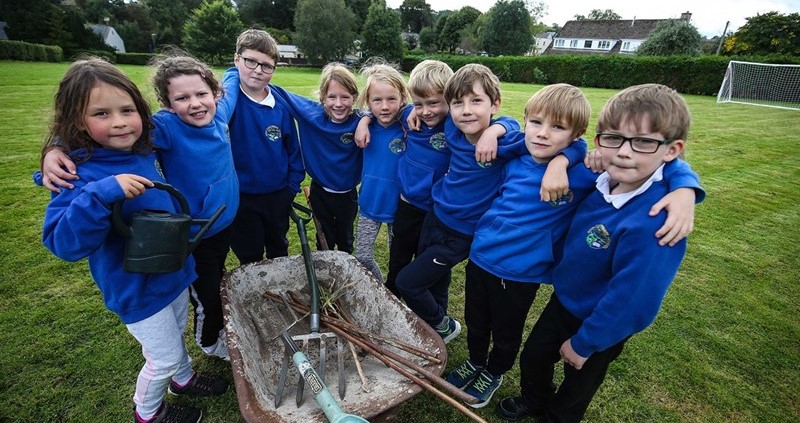 A photo of nine primary school children huddled around a wheelbarrow filled with garden equipment, such as a rake, spade and watering can.
