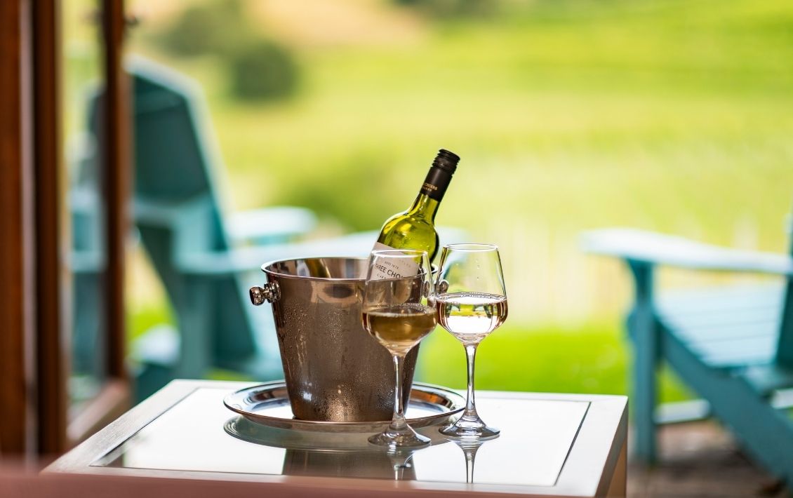 A bottle of wine in a wine cooler and wine glasses on a table overlooking a lawn 